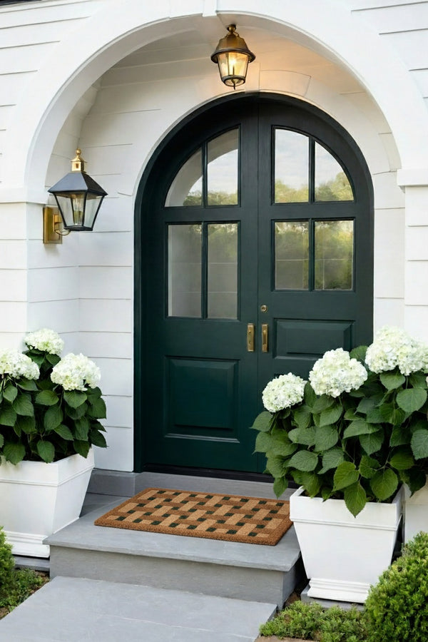 Green front door with white flowers and a doormat on a white house exterior.